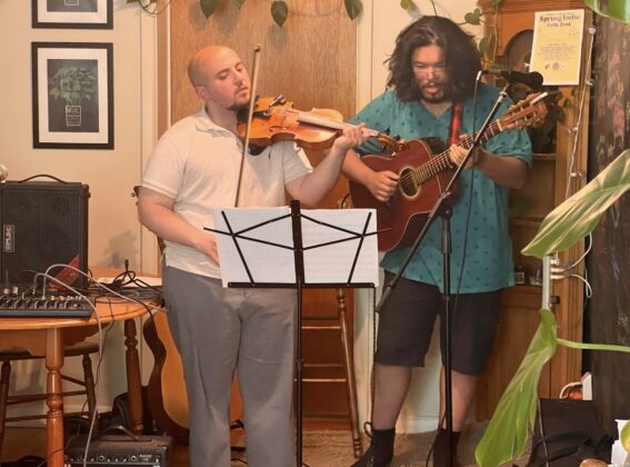 One person with a violin and another with a guitar stand in front of music sheets and perform in a house.